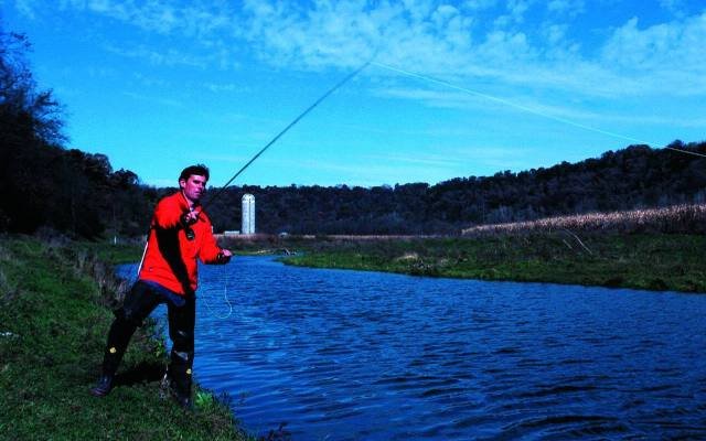 An angler casting a fly rod on a river under a blue sky