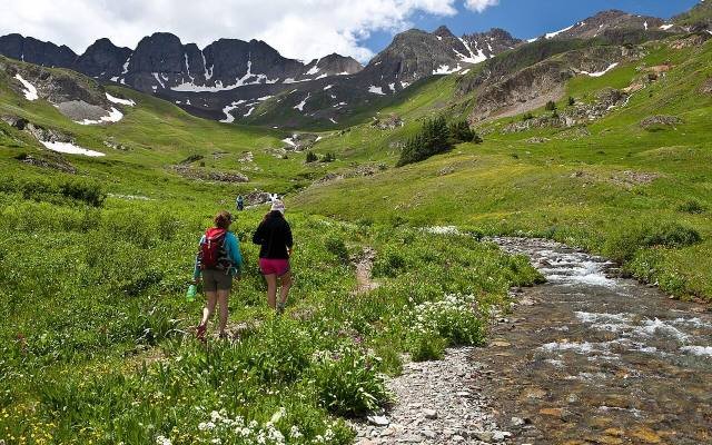 Two hikers walking beside a creek in a green alpine valley below jagged peaks